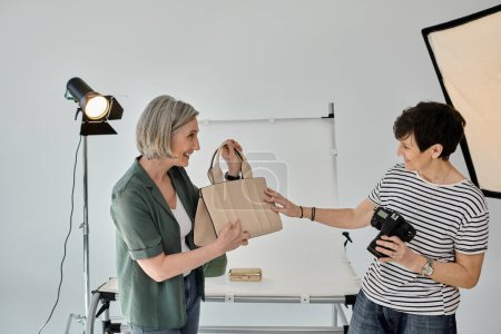 A middle-aged lesbian couple in a professional photo studio, one holding a bag and the other holding a camera.