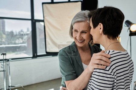 Lesbisches Paar mittleren Alters umarmt sich herzlich in einem modernen Fotostudio und fördert ein Gefühl der Zusammenarbeit und Einheit.