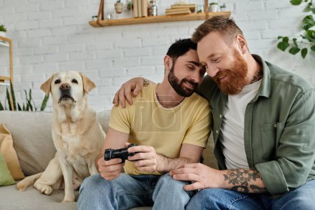 Two men, engrossed in a video game, sit on a couch in their living room with their dog by their side.