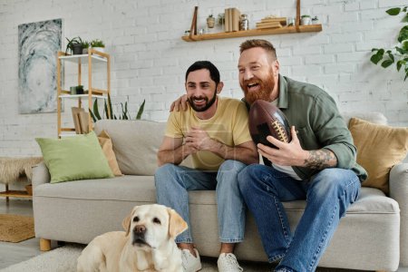 Un homme avec une barbe est assis sur un canapé à côté d'un Labrador, se détendre et regarder un match de sport.