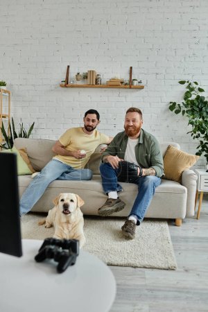 Two bearded men relax on a couch with their Labrador dog, watching a baseball match on TV in a cozy living room.