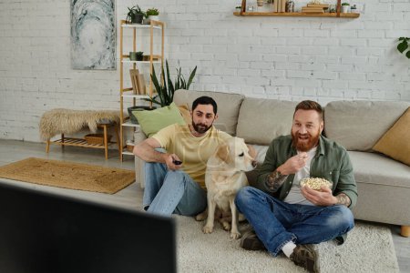 Photo for Two men, one with a beard, sitting on the floor, watching movie with their Labrador dog, eating popcorn. - Royalty Free Image
