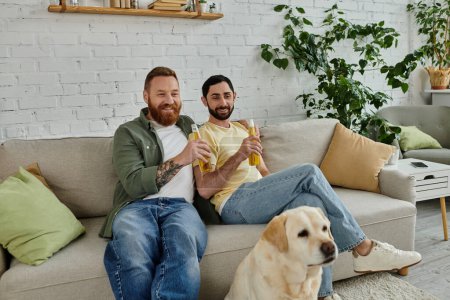 Zwei Männer, ein schwules Paar, sitzen mit ihrem Hund auf einer Couch und verfolgen gemeinsam im Wohnzimmer ein Sportspiel..