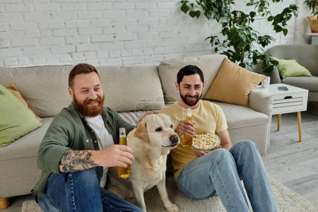 Two bearded men happily watch a sports match with their labrador dog on the couch in the living room.