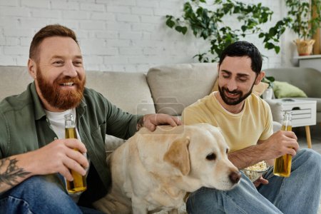 Two men, a gay couple, relax on a couch watching a sports match with their Labrador dog in their living room.