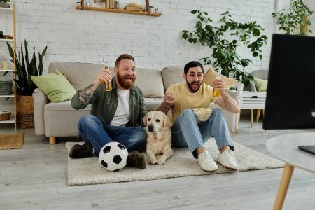 Two men, with beards, sit on rug with Labrador dog, watching sport match in living room.