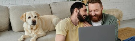 Bearded gay couple and their Labrador dog sitting on a couch, both engrossed in viewing content on a laptop.