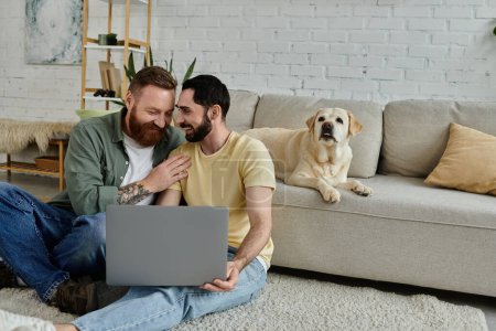 Two men, a bearded gay couple, sit on the floor with a laptop, working remotely in their living room near a labrador dog.