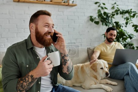 Ein bärtiger Mann sitzt auf einer Couch, telefoniert mit einem Handy und arbeitet in einem gemütlichen Wohnzimmer neben seiner Partnerin und einem Labrador-Hund..