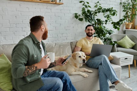 Foto de Bearded gay couple sits on a couch with a labrador dog, working remotely on a laptop in a cozy living room. - Imagen libre de derechos
