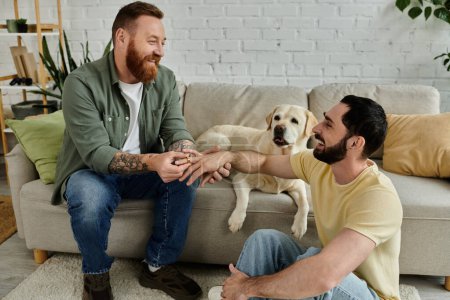 Two men with beards, sit on a couch with labrador dog in a cozy living room setting, marriage proposal