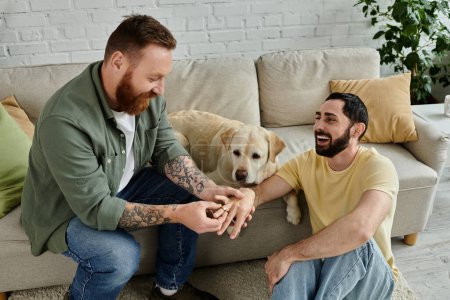 Foto de Bearded gay man sitting on a couch next to a labrador dog, making marriage proposal to partner - Imagen libre de derechos