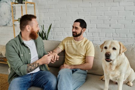 Two men with beards sit on a couch next to their Labrador dog in a cozy living room, marriage proposal