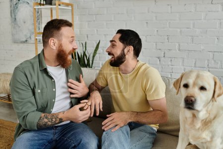 Bearded gay couple sits on couch with labrador dog, marriage proposal