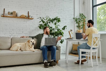 Ein bärtiger Schwuler sitzt auf einer Couch mit Laptop neben einem Labrador-Hund in einem gemütlichen Wohnzimmer und arbeitet aus der Ferne.