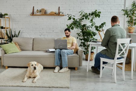 A bearded man works on a laptop while sitting on a couch next to a relaxed labrador dog in a cozy living room setting.