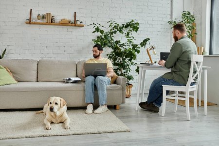 Bearded gay man sits on couch with laptop, working beside labrador dog in cozy living room setting.