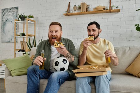Foto de Two men with beards sitting on a couch, enjoying pizza and beer with a labrador dog in the living room. - Imagen libre de derechos