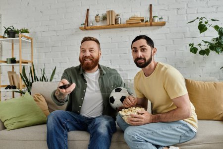 Foto de Two bearded men sit on a couch holding a soccer ball, enjoying quality time together in their cozy living room. - Imagen libre de derechos