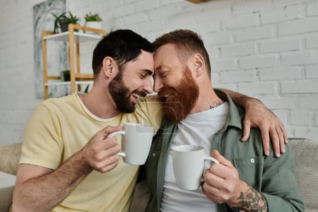 Two men with beards sit on a couch, holding coffee mugs, in a cozy living room.