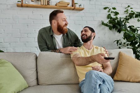 A bearded gay couple sits on a couch, holding a remote, watching television