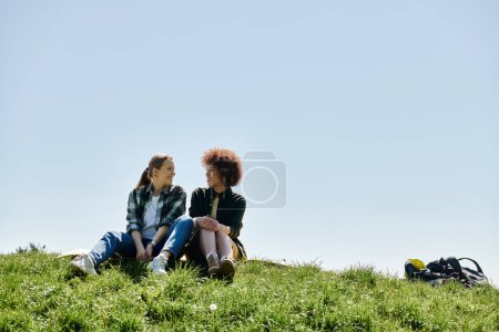 Zwei junge Frauen, eine mit braunen und eine mit lockigen dunklen Haaren, sitzen auf einem grasbewachsenen Hügel und genießen ihre Wanderung.