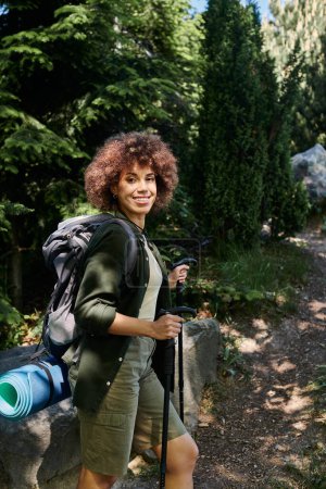 A woman smiles as she hikes through a lush green forest, her backpack and hiking poles by her side.