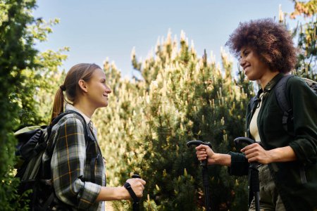 Dos mujeres jóvenes, una con un afro y la otra con el pelo liso, caminan por un bosque en un día soleado, sonriendo y charlando.