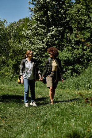Zwei junge Frauen, eine weiße und eine schwarze, gehen Hand in Hand durch einen sattgrünen Wald und genießen eine gemeinsame Wanderung..