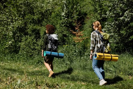 Two young women, a lesbian couple, hike through a green forest with backpacks and rolled-up sleeping mats.