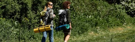 Two young women, one with long blonde hair and the other with short afro hair, hike through a wooded trail, carrying backpacks and rolled-up sleeping mats.