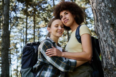 Zwei junge Frauen, ein lesbisches Paar, stehen zusammen in einem Wald und umarmen sich. Beide tragen Rucksäcke und genießen die Wildnis.