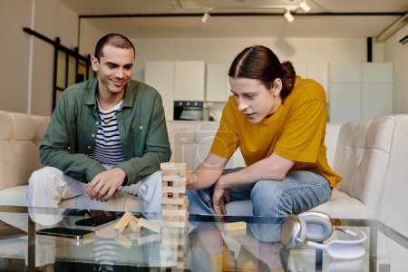 Two young men, dressed casually, play a game of blocks in their modern apartment.