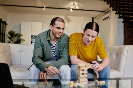 Un jeune couple gay profite d'une soirée détendue à la maison, jouant à un jeu de blocs et riant ensemble sur un canapé blanc dans un appartement moderne.