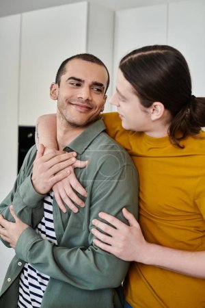 Two young men, dressed casually, share a tender moment in their modern apartment.