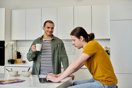 A young gay couple relaxes in their modern apartment, one holding a mug of coffee while the other works on a laptop.