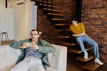 Un jeune couple gay se détend dans leur appartement moderne, l'un sur le canapé défilant sur leur téléphone, l'autre dans les escaliers à l'aide d'un ordinateur portable.