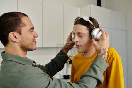 A young gay couple relaxes in their modern apartment, one putting headphones on the other.