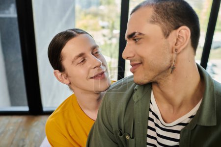Two young gay men in casual attire share a loving look in a modern apartment.