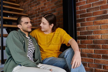 Ein junges schwules Paar sitzt zusammen in einer modernen Wohnung, die Augen in einem zärtlichen Moment der Zuneigung verschlossen.