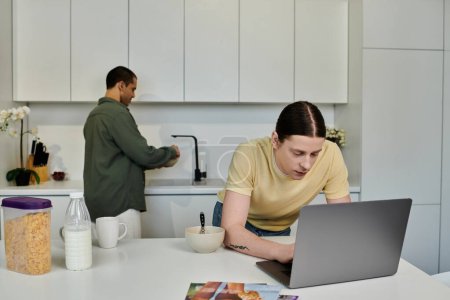 Un jeune homme travaille sur son ordinateur portable pendant que son partenaire lave la vaisselle en arrière-plan. Ils sont dans une cuisine moderne appartement avec armoires blanches et un comptoir blanc.