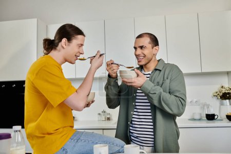 Two young men in casual attire share a lighthearted moment while eating breakfast in their modern kitchen.