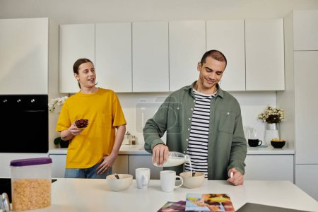 A young gay couple enjoys breakfast together in their modern apartment. One man pours milk into a bowl while the other watches with a smile.