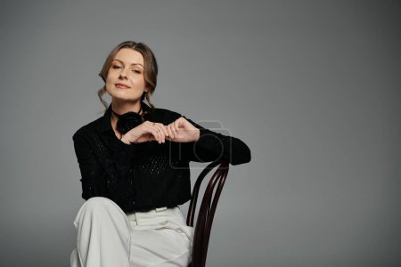 Una mujer con una blusa negra y pantalones blancos se sienta en una silla, mirando con confianza a la cámara.