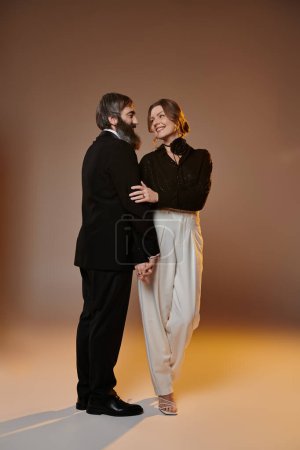 Ein Paar posiert gemeinsam in einem Studio-Setting, in eleganter Kleidung, wobei die Frau den Mann liebevoll anlächelt.