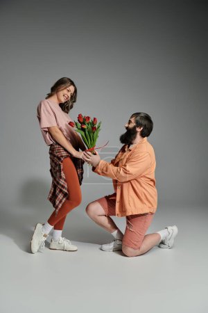 A man in a peach shirt and shorts proposes to a woman in a pink shirt and brown leggings, holding a bouquet of tulips.