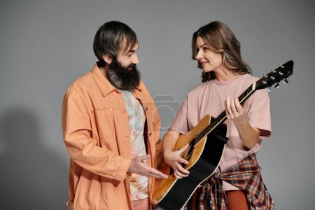 A couple in stylish attire share a musical moment, with one playing a guitar while the other looks on lovingly.
