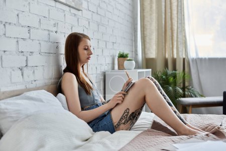 Eine junge Frau mit Tätowierungen sitzt auf ihrem Bett in einer modernen Wohnung und schreibt in ein Notizbuch.