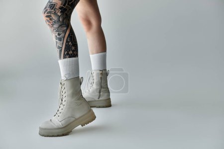 A close-up of a tattooed womans legs, showcasing white boots and white socks against a gray backdrop.