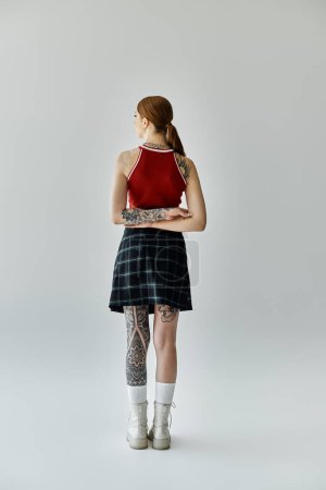 Une jeune femme tatouée portant une jupe à carreaux et un débardeur rouge se tient le dos à la caméra, regardant par-dessus son épaule.